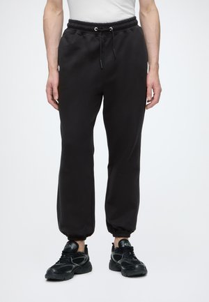 Person trägt schlichte schwarze Jogger-Hosen mit elastischen Bündchen und Kordelzug, kombiniert mit schwarzen Sportschuhen vor weißem Hintergrund.