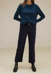 Marinblå stickad tröja med axelknappdetalj, tillsammans med högmidjade vida byxor. Modellen bär svarta ankelstövlar.