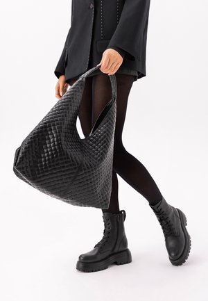 Isik mustas riietuses hoiab suurt musta kootud käekotti, kannab musti sukkpükse ja massiivseid musta paeltega saapaid, kõnnib valgel taustal.