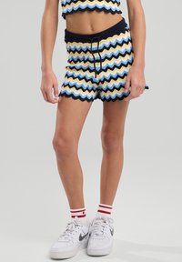 Shortsit, joissa on aaltoileva raidallinen kuosi sinisessä, keltaisessa ja mustassa. Kairahemmo, nauhavyötärö, yhdistetty valkoisten tennareiden ja raidallisten sukkien kanssa.