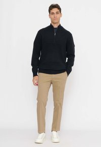 Uomo che indossa un maglione nero a mezzo zip, pantaloni beige e scarpe da ginnastica bianche, in piedi con una mano in tasca davanti a uno sfondo bianco semplice.