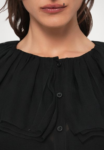 Čierna blúzka s plisovaným, volánikovým výstrihom, s gombíkovým zapínaním a textúrovanou tkaninou. Detailný záber na vrchný odev bez úplného pohľadu.