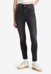 Mujer vistiendo jeans skinny de tiro alto en gris oscuro, top corto blanco, calcetines blancos y zapatillas crema con suelas de goma sobre fondo blanco.