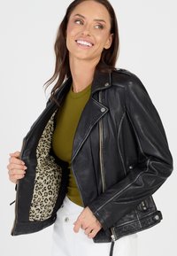 Schwarze Leder-Motorradjacke mit silberner Hardware, Reißverschluss-Taschen und einem Leo-Muster-Futter, getragen über einem olivgrünen Oberteil.