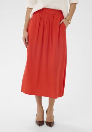 Femme portant une jupe midi rouge vif taille haute à plis, associée à des escarpins marron pointus et un pull en maille blanc.