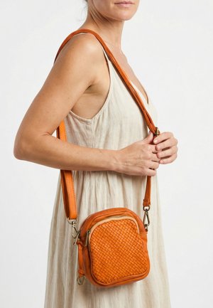 Vrouw in een beige mouwloze jurk met een kleine oranje geweven leren crossbodytas met verstelbare riem.