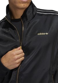 Svart satinträningsjacka med gulddragkedja, tre vita ränder på axlarna och en präglad guld Adidas-logotyp på bröstet.