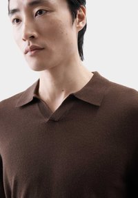 Bruine V-halstrui van zacht breisel, met een eenvoudige kraag en een gladde textuur, gedragen door een model met kort haar.