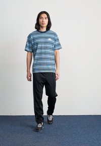 Stribet blå t-shirt med korte ærmer og rund hals, parret med sorte bukser og træningssko, stående mod en ensfarvet baggrund.