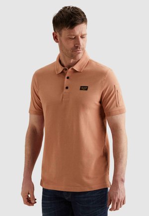 Mann trägt ein hellbraunes kurzärmliges Poloshirt mit einem kleinen schwarzen Logo-Aufnäher auf der Brust und dunkelblaue Jeans.