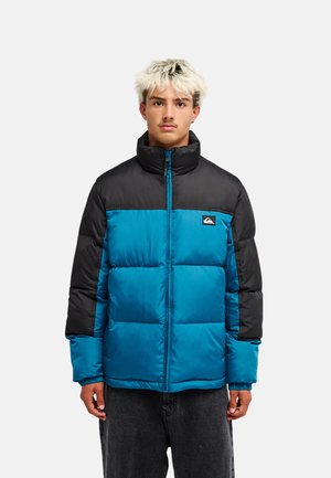 Junger Mann mit blondem Haar, der eine blau-schwarze Daunenjacke trägt und vor einem schlichten hellen Hintergrund steht.