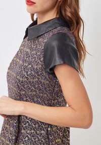 Vestido con una tela estampada en púrpura y dorado, con una cintura estructurada, mangas cortas de cuero negro y un diseño de cuello clásico.