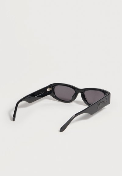 Lunettes de soleil rectangulaires noires avec des verres foncés et « Jumpsuit » embossé sur des branches larges, présentées sur un fond clair uni.