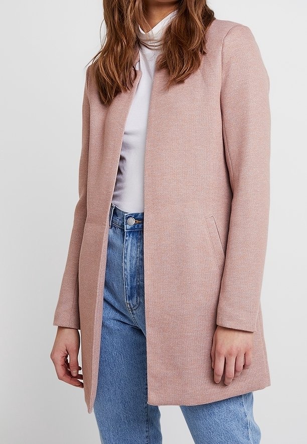 Femme portant un manteau rose clair jusqu'aux genoux par-dessus un col roulé blanc et un jean bleu, debout devant un fond uni.