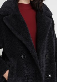 Abrigo negro de doble botonadura hecho de tela texturizada, con grandes botones negros y solapas anchas, llevado sobre una blusa burdeos.