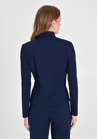 Veste tailleur bleu marine avec un col haut, une coupe ajustée, des manches longues et une texture lisse. Comprend des coutures subtiles et des fentes au dos.