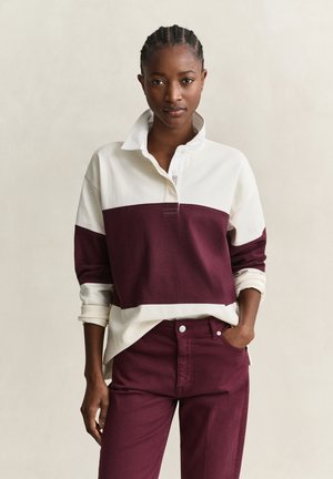 Femme aux cheveux tressés portant un maillot de rugby à manches longues avec col blanc et bordeaux et un pantalon bordeaux assorti, debout avec une main dans la poche.
