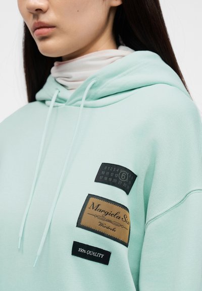 Φούτερ με κουκούλα σε μέντα πράσινο χρώμα, με πολλαπλές μαύρες και καφέ υφασμάτινες ετικέτες με κείμενο. Μαλακή υφή, ρυθμιζόμενη κουκούλα και κορδόνια για εφαρμογή.