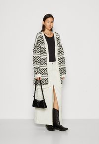 Cárdigan de gran tamaño tejido en negro y blanco con patrones geométricos, usado sobre una blusa negra, combinado con una falda beige y botas negras.