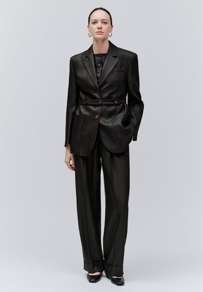 Femme portant un costume noir sur mesure avec un blazer ceinturé, un pantalon à jambes larges, des boucles d'oreilles créoles dorées et des chaussures à talons noires, debout devant un fond gris.