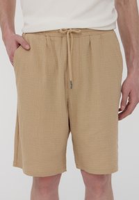 Bermuda beige, con texture, dalla vestibilità ampia con vita elasticizzata e cordoncino. Presenta tasche laterali e un orlo leggermente sfilacciato.