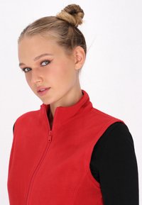 Vesta din fleece roșu, cu guler înalt și fermoar în față. Textură netedă, design fără mâneci, purtată peste un top negru cu mânecă lungă.