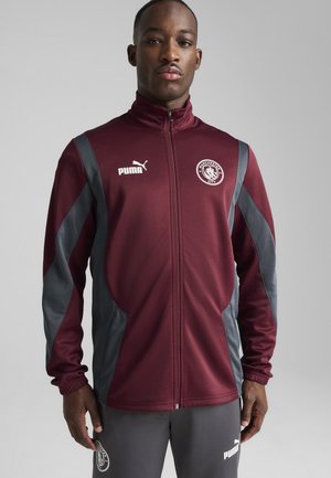 Chaqueta deportiva marrón con acentos grises, cierre con cremallera, cuello alto y logotipo en el pecho. Tejido suave con diseño ajustado y puños acanalados.
