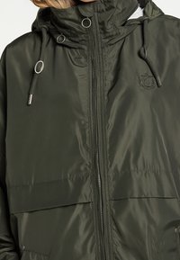 Chaqueta impermeable de color verde oscuro con cuello alto, capucha ajustable, cierre de cremallera y detalles metálicos. Cuenta con un logo en el lado izquierdo.