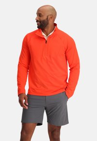 Oranje lange mouwen kwart-rits shirt met een borstzak, gecombineerd met grijze shorts. Het shirt heeft een gestructureerde stof en ribgebreide manchetten.