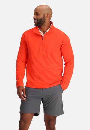 Oranges Langarm-Shirt mit Viertelreißverschluss und einer Brusttasche, kombiniert mit grauen Shorts. Das Shirt hat einen strukturierten Stoff und gerippte Bündchen.