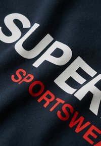 Marineblåt bomuldsstof med iøjnefaldende hvid tekst "SUPER" og rød tekst "SPORTSWEAR". Materialet har et tekstureret, vævet udseende.