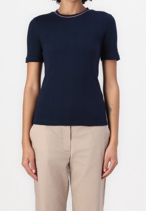 Femme portant un haut côtelé bleu marine à manches courtes avec une discrète bordure rouge et blanche autour du col rond, et un pantalon beige.