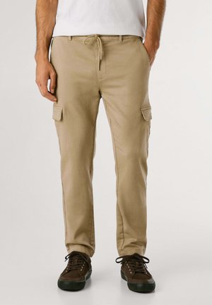 Khaki cargobroek met twee zijzakken, elastische tailleband met trekkoord en taps toelopende pijpen. G gedragen met donkerbruine sneakers.