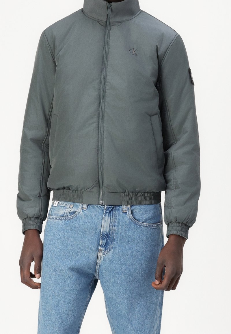 Graue Nylon-Bomberjacke mit hohem Kragen, Frontreißverschluss, elastischen Bündchen und Saum; verfügt über ein kleines Logo auf der Brust.