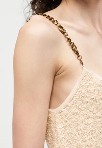 Close-up of a person wearing a textured beige sleeveless top with a gold chain strap on the shoulder.