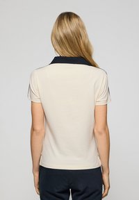 Camisa polo beige con cuello azul marino y mangas cortas. Tejido suave con aberturas laterales. Diseño sencillo, apto para uso casual.