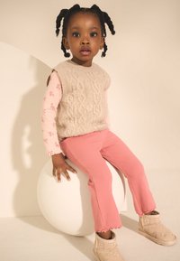 Bēša adīta bezroku džemperis pār sārtirozā ziedu raksta garroku kreklu, kopā ar sārtiem korduroja biksēm un gaiši bēžām zābakiem uz baltas sfēras.