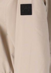 Tissu de veste beige avec une texture lisse ; présente un logo carré noir avec un graphique de montagne sur la manche.