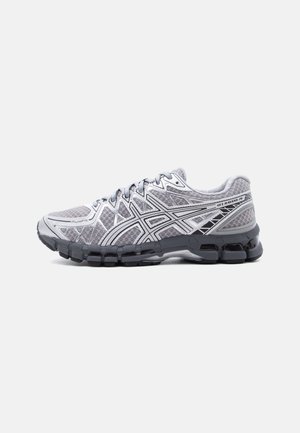 Zapatilla de correr gris claro con suela negra, parte superior de malla, franjas laterales geométricas en blanco y negro, y texto "GEL-KAYANO 28" cerca del talón.