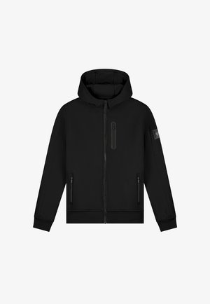 Blonder Junge steht, trägt einen schwarzen Kapuzenpullover mit Reißverschluss, ein weißes T-Shirt, hellblaue zerrissene Jeans und schwarz-weiße Turnschuhe vor einem schlichten Hintergrund.