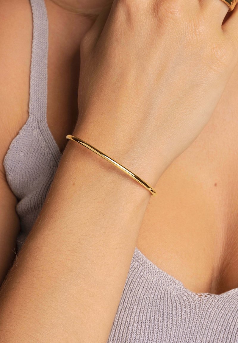 Gros plan d'un avant-bras portant un bracelet fin et lisse en or avec un haut en maille lavande visible en arrière-plan.