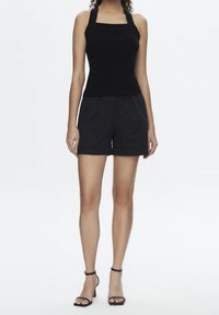 Zwarte aansluitende tanktop met vierkante halslijn, gecombineerd met hoge zwarte shorts met plooien en opgerolde zoom. Zwarte sandalen met straps.