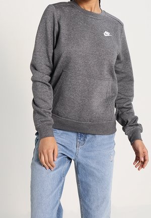 Personne portant un sweat-shirt gris Nike avec poche avant et un jean bleu clair, debout devant un fond clair uni.