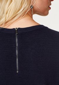 Ljusblond person som bär en ribbad marinblå tröja med en synlig silverdragkedja på ryggen och en svart och silver hängande örhänge.