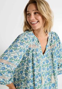 Blouse à motif floral en tissu léger avec des fleurs bleues et des vignes vertes. Elle a un décolleté en V, des accents en dentelle et des liens à frange au niveau du col.