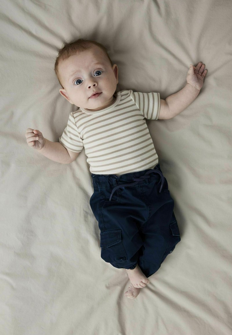 Bebe v pruhovaném béžovo-bílém body doplněném o námořnické cargo kalhoty. Měkké, neutrální pozadí s dítětem ležícím na lehké látce.