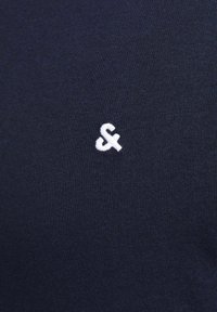 Navy blue fabric with a smooth texture features a small, white embroidered logo resembling an "F" positioned on the left side.