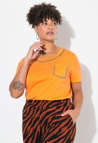 Lys orange T-shirt med rund halsudskæring, prydet med sølvfarvede nitter omkring kraven og en lomme. Kombineret med sort og orange stribede bukser.