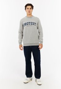 Grijze sweatshirt met "PROTEST" en "SPORTSWEAR" in marineblauw gedrukt. Heeft een ronde halslijn en geribbelde manchetten. Gepaard met donkere jeans en sneakers.