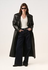 Czarny skórzany płaszcz trench, biały top z dekoltem w serek, szerokie ciemnoniebieskie dżinsy i czarne buty. Model noszący czarne okulary przeciwsłoneczne, ręce w kieszeniach.